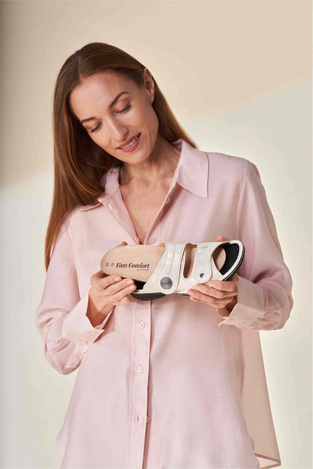 Donna sorridente con una camicia rosa tiene in mano una ciabatta FinnComfort Sansibar color sabbia dall’ampia base d’appoggio per il comfort e la stabilità. Dalla ciabatta Sansibar, la donna sta estrendo il plantare FinnComfort.