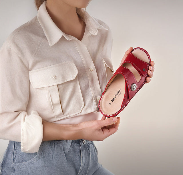 in primo piano una figura femminile con in mano una ciabatta donna FinnComfort di colore rosso. Il modello della ciabatta si chiama Moorea.