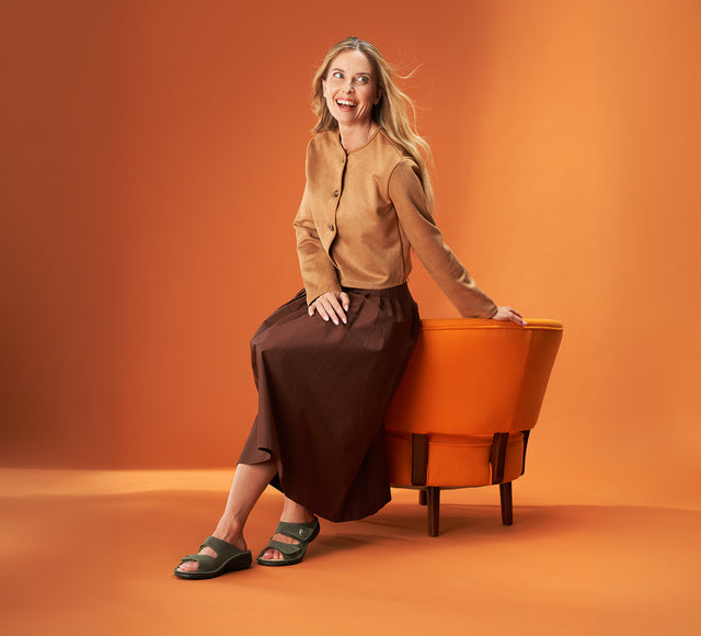 donna sorridente appoggiata al retro di una poltrona arancione indossando ciabatte finncomfort colina verdi abbinate a gonna ampia marrone e camicia beige