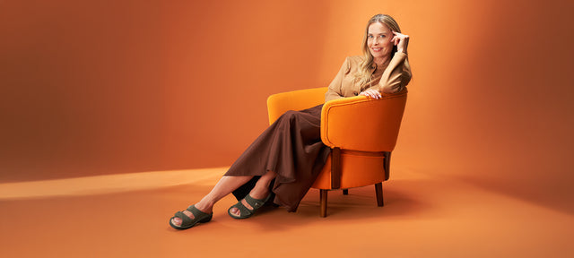 donna sorridente seduta su poltrona arancione vestita con gonna ampia marrone e camicia beige abbinati a colina verde ciabatte finncomfort